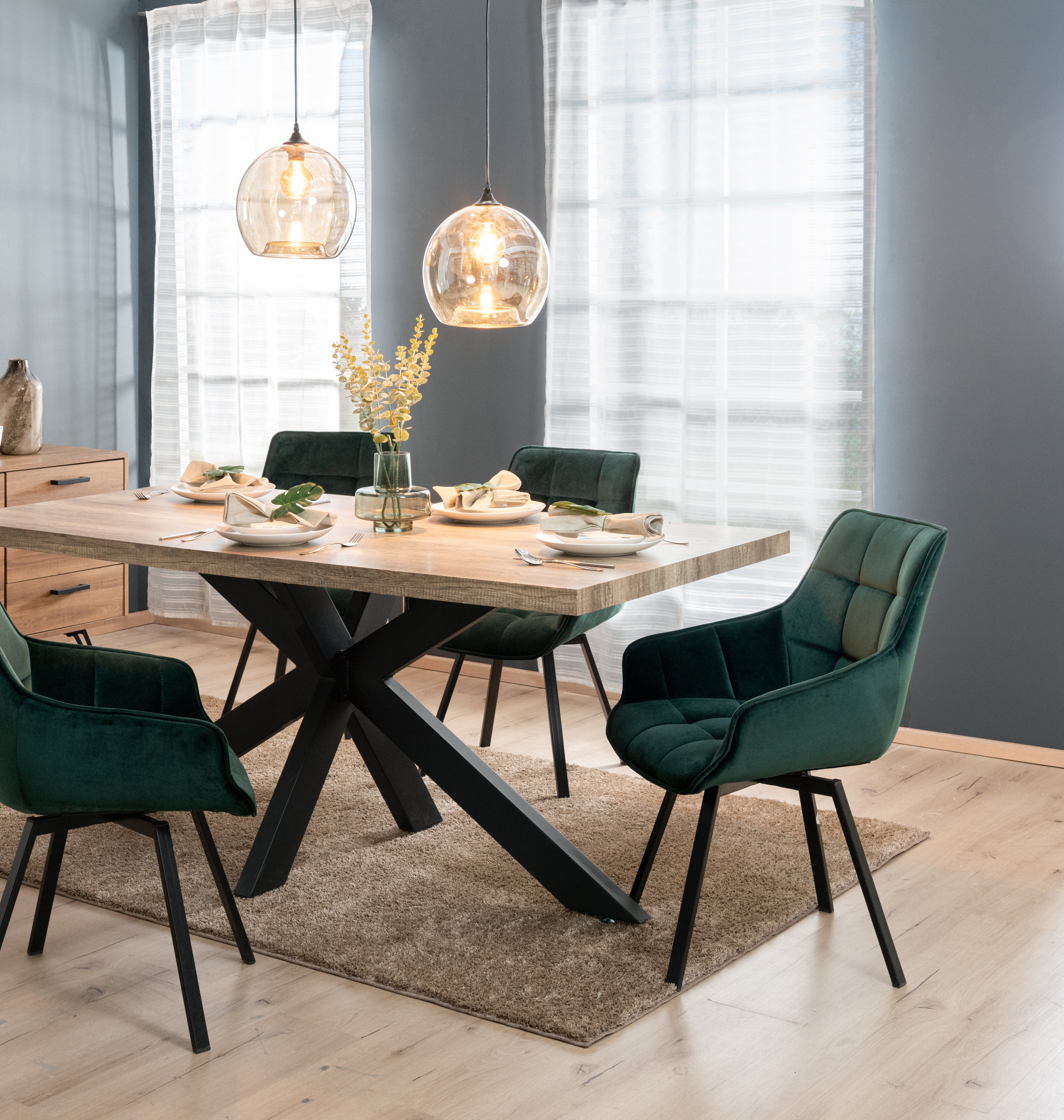 Modern dining room with rustic wooden table, green velvet chairs, and hanging pendant lights, creating a cozy, stylish ambiance.