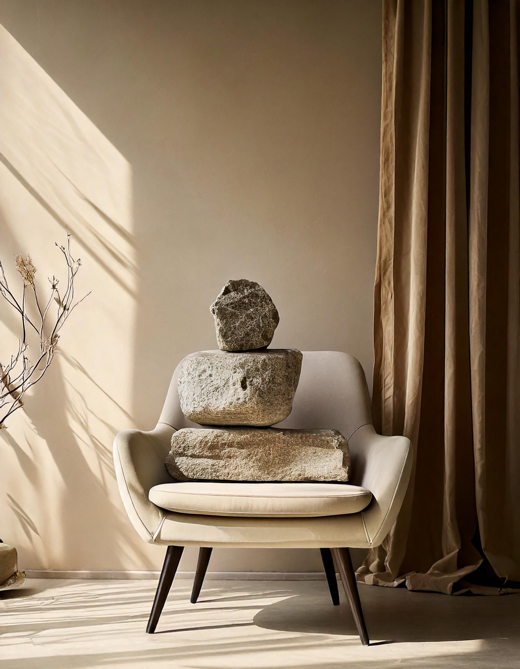 Cozy interior with a beige armchair and stacked stones, alongside a soft curtain and dried branch in natural sunlight.