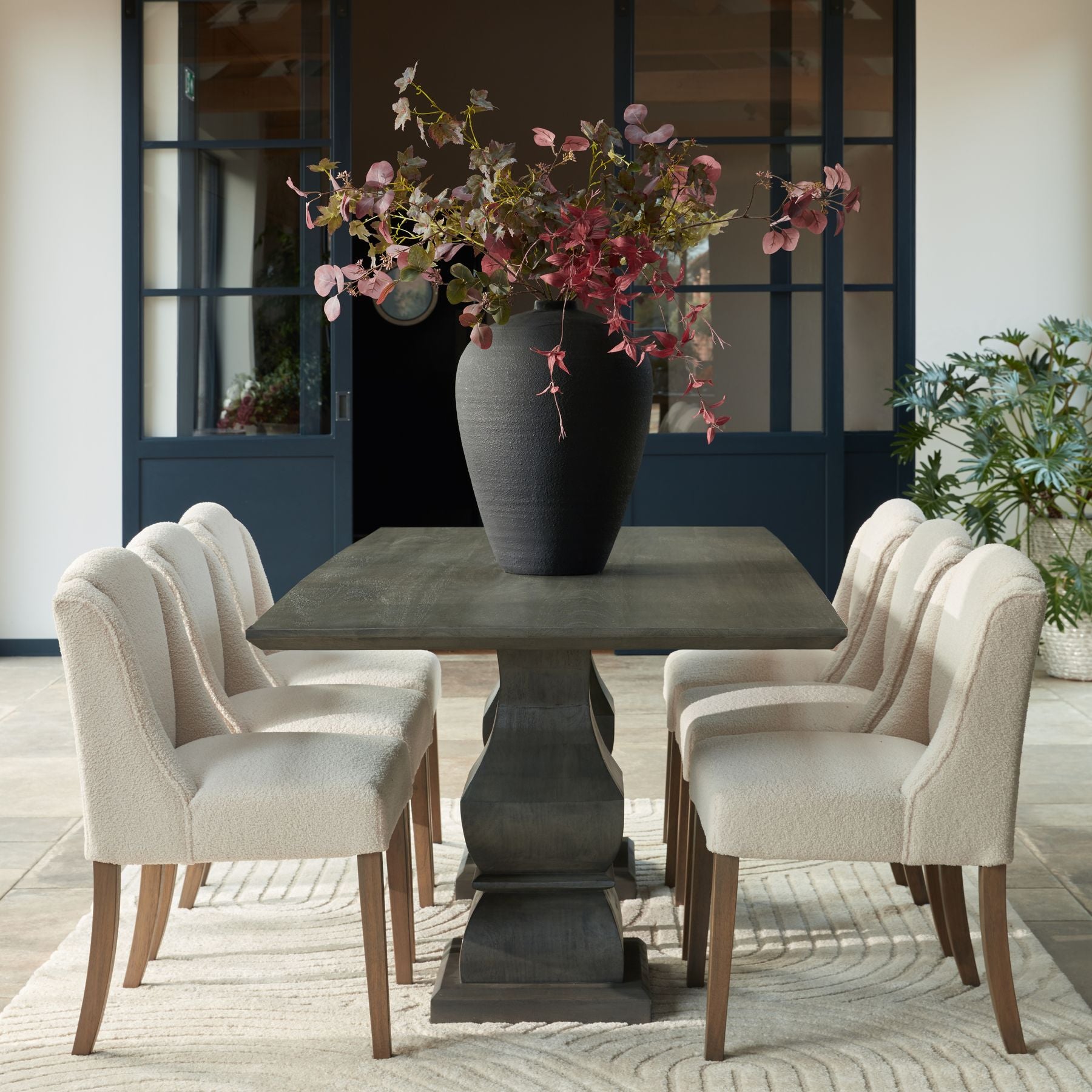 Elegant dining area with a dark wooden table, six upholstered chairs, and a large vase with pink foliage, set against glass-paneled doors.