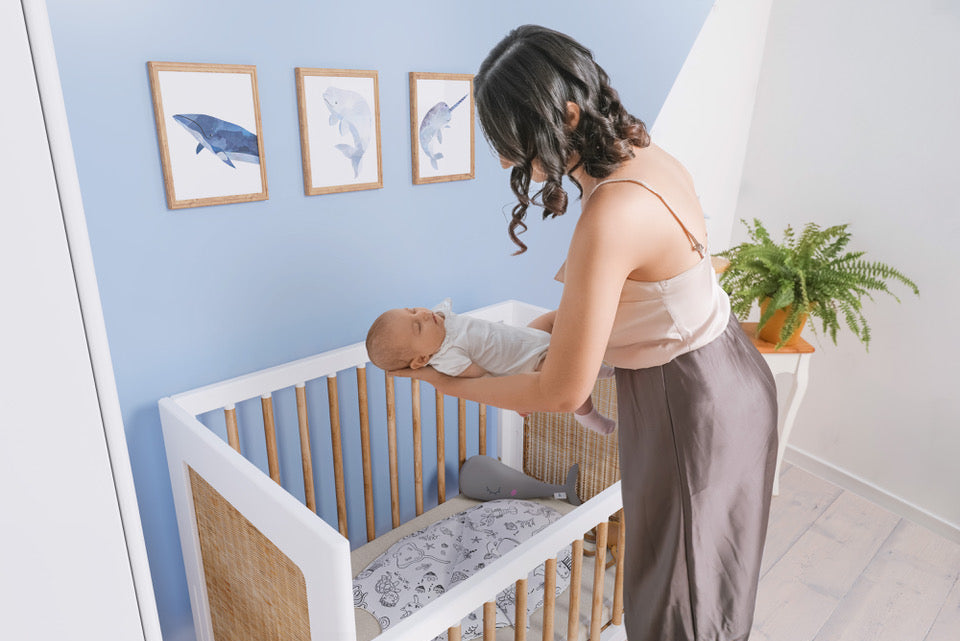 Mother gently placing baby in a crib with blue painted wall and framed dolphin artwork, next to a fern plant.