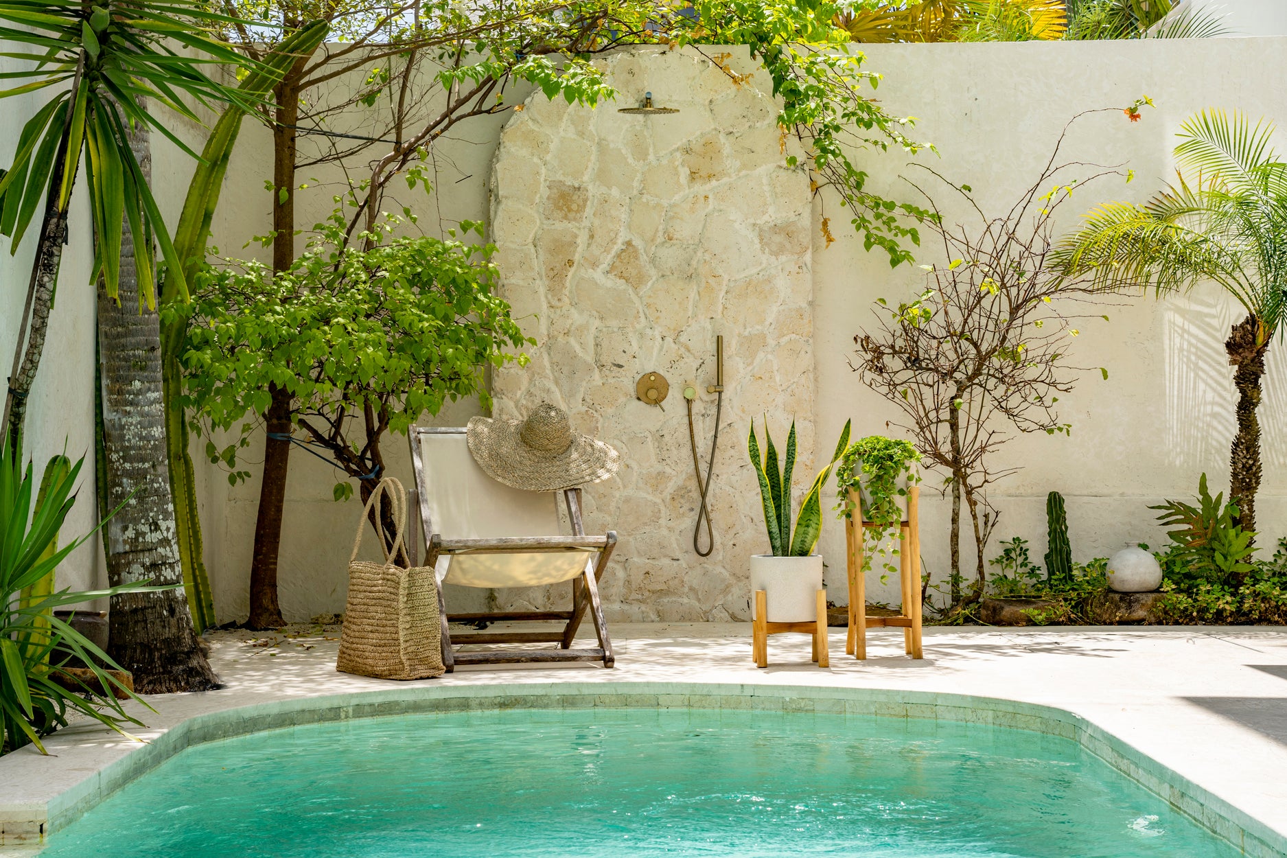 Serene poolside patio with lush greenery and a rustic stone shower wall, complemented by a straw chair and woven basket.
