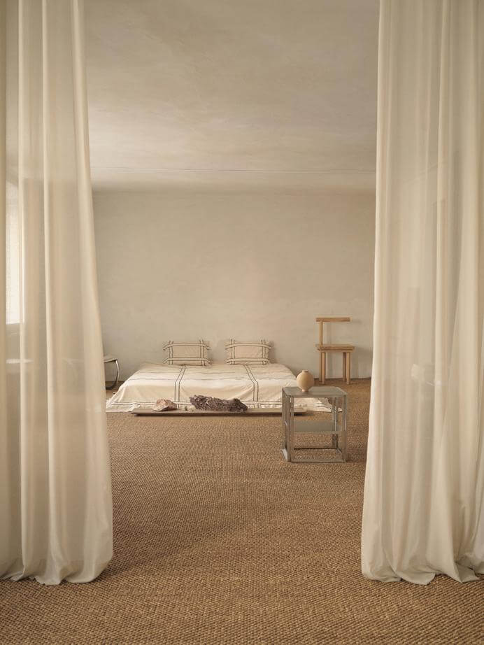 Minimalist bedroom with sheer curtains, neutral tones, and modern furniture, featuring a low-profile bed and woven rug.